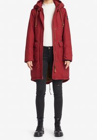 Rode parka met een capuchon, trekkoordaccenten en twee voorzakken. Gedragen over een witte sweater en zwarte skinny jeans, gecombineerd met zwarte laarzen.