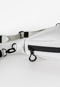 Weißer Stoff-Crossbody-Beutel mit schwarzem Reißverschluss, ausgestattet mit einem Nylonriemen, Metallbeschlägen und einer dreieckigen Form.