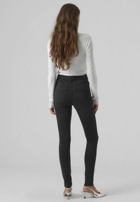 Schwarze hoch taillierte Skinny-Jeans mit glatter Textur und zwei Gesäßtaschen, kombiniert mit einem weißen Langarmoberteil und silbernen Absätzen.
