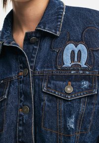 Tumman pesun farkkutakki, jossa on brodeerattu Mikki Hiiri -kuvio. Ominaisuudet: klassinen kaulus, napillinen etuosa ja kuluneet yksityiskohdat.