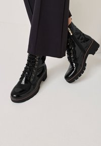 Bottines en cuir verni noir avec un laçage à l'avant et une fermeture éclair sur le côté, semelle épaisse avec un profil texturé, dotées d'œillets dorés.