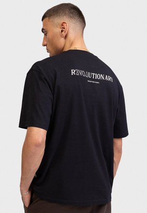 Tânăr cu fața spre stânga, purtând un tricou negru cu textele „REVOLUTIONARY” și „REDEFINED REBEL” pe partea superioară a spatelui, pe un fundal simplu.