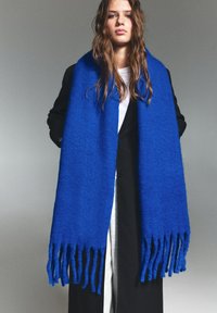 Blå uld tørklæde med frynser, tyk tekstur, rektangulær form, båret over en sort frakke og en hvid skjorte.