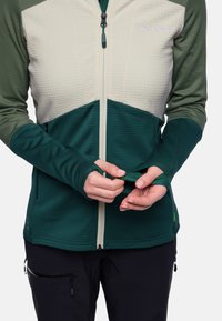 Fleecejacke mit einem Reißverschluss an der Vorderseite, mit einem cremefarbenen oberen und einem dunkelgrünen unteren Bereich. Enthält Daumenlöcher und strukturierte Stoffakzente.