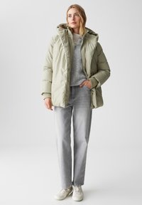Giacca a vento verde chiaro con cappuccio, dotata di chiusura con zip e bottoni a pressione. Indossata sopra un maglione grigio e abbinata a jeans grigi chiari e sneakers bianche.