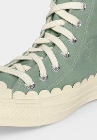 Υψηλό αθλητικό παπούτσι με ανοιχτοπράσινο ύφασμα, κρεμ χρωματιστή λουλουδωτή λαστιχένια σόλα και λευκά κορδόνια που περνούν μέσα από μεταλλικές οπές.