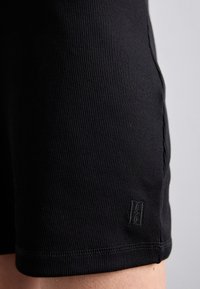 Gros plan d'un vêtement en tissu côtelé noir avec un petit logo brodé près de l'ourlet porté sur la jambe d'une personne.