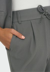 Szare spodni o dopasowanym kroju z prążkowanej tkaniny, elastycznym pasem i sznurkiem do wiązania. Posiada boczne kieszenie i delikatne fałdy.