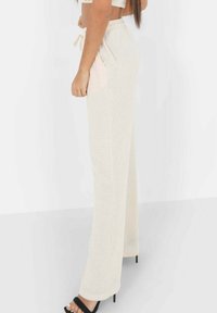 Pantalons de détente en tricot beige avec une coupe ample, taille élastique à cordon, poches latérales et texture douce. Les pantalons sont assortis à des talons noirs.