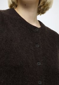 Tmavohnedý sveter na zapínanie na gombíky, vyrobený z huňatej látky, s okrúhlym výstrihom a piatimi čiernymi gombíkmi po celej prednej strane. Mäkká textúra, minimalistický dizajn.