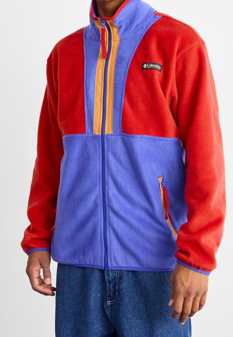 Chaqueta de forro polar roja y morada con un alto cuello. Presenta cierre de cremallera, acentos naranjas y un pequeño logo. Detallada con bolsillos laterales.