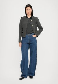 Vrouw met lang zwart haar, gekleed in een grijze knopenjas, een zwart-witte gestreepte shirt, wijde blauwe jeans en zwarte schoenen, staande met haar hand in de zak.
