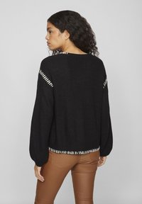 Čierny pletený sveter s balónovými rukávmi, s bielymi stehmi pozdĺž ramien a vzorovaným lemom. Nosený cez hnedé nohavice.