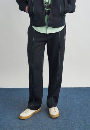 Oseba, oblečena v črne hlače z modrimi vertikalnimi šivi, svetlo zeleno srajco, črno jakno in bele superge, stoji na modrem preprogi.