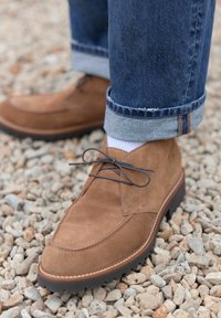 Cizme de gleznă din suede maro, cu vârful rotund, design cu șireturi și talpă texturată, asortate cu blugi denim suflecați și șosete albe.