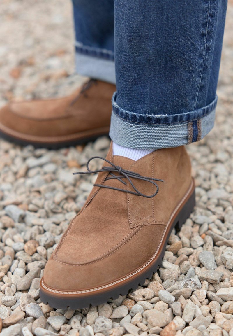 Cizme de gleznă din suede maro, cu vârful rotund, design cu șireturi și talpă texturată, asortate cu blugi denim suflecați și șosete albe.