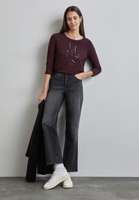 Burgunderfarbenes Langarmhemd mit geprägtem Text, kombiniert mit hochgeschnittenen schwarzen Jeans und weißen Sneakers. Strukturierter Stoff und schlichte Form.