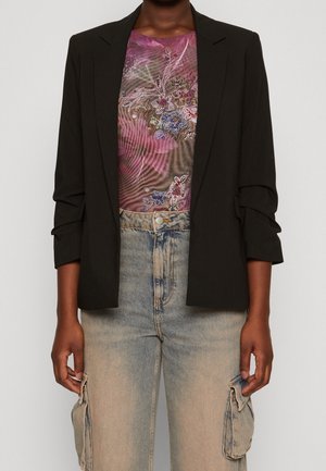 Person trägt ein schwarzes Sakko, ein lila, mit Blumen besticktes Oberteil und ausgewaschene beige Cargo-Jeans mit Seitentaschen, steht vor einem schlichten Hintergrund.