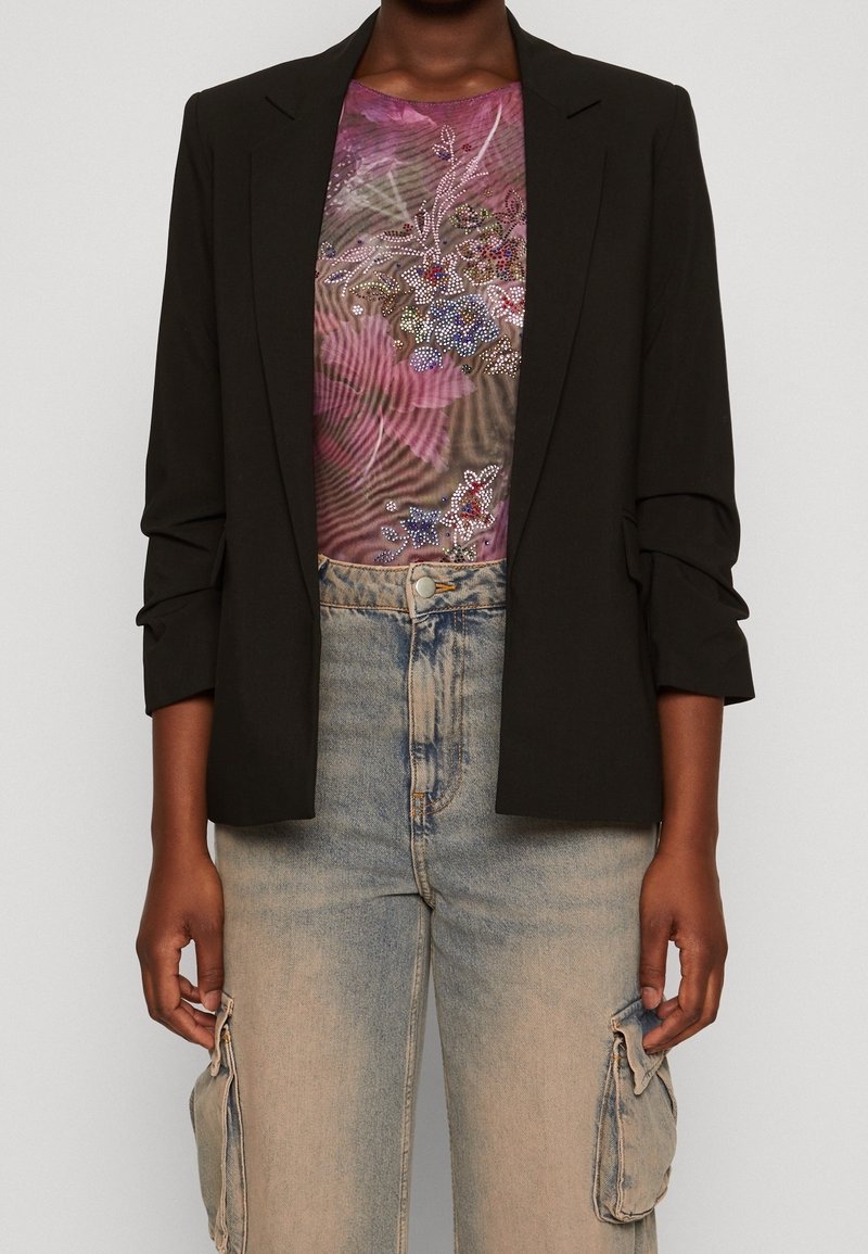 Personne portant un blazer noir, un haut brodé de fleurs violettes, et un jean cargo beige délavé avec poches latérales, debout devant un fond uni.