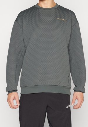 Personne portant un sweat-shirt gris texturé à manches longues et col rond avec un petit logo "TERREX" sur la poitrine et un pantalon noir avec un logo sur la cuisse.