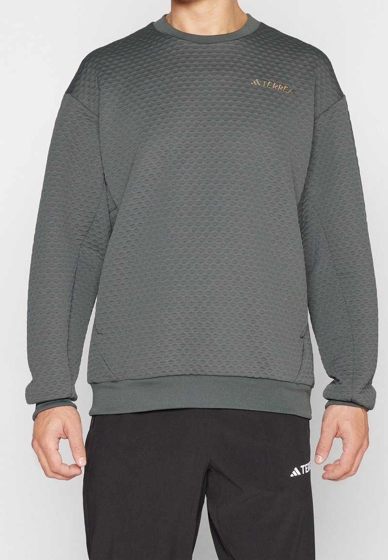 Personne portant un sweat-shirt gris texturé à manches longues et col rond avec un petit logo "TERREX" sur la poitrine et un pantalon noir avec un logo sur la cuisse.