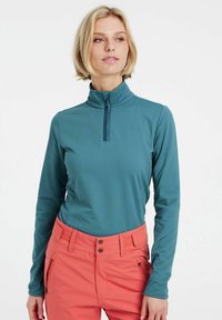 Maglia a maniche lunghe color teal con colletto alto e zip a metà, realizzata in un tessuto liscio e leggero; abbinata a pantaloni da sci rosa.