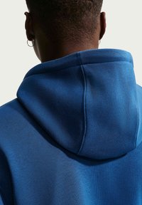 Nahaufnahme einer Person, die eine blaue Kapuzenjacke trägt, von hinten gezeigt, mit kurzen Haaren und einem kleinen Creolenohrring im linken Ohr.