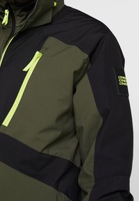 Veste vert foncé et noire avec des fermetures éclair jaune néon et un patch rectangulaire sur la partie supérieure de la manche marqué plusieurs fois du texte "ONE".
