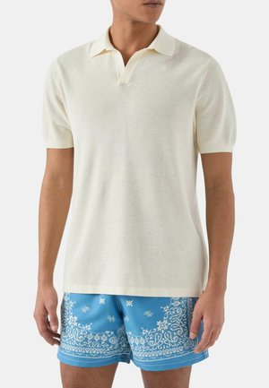 Uomo che indossa una polo bianca in maglia a maniche corte e pantaloncini da bagno blu con fantasia, in piedi davanti a uno sfondo semplice.