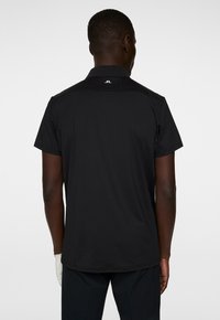 Svart kortärmad polotröja med krage, med en texturerad rygg och en vit logotyp vid nacken. Slätt tyg, vanlig passform.