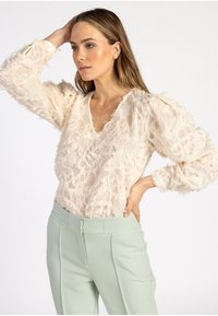 Beige textuurblouse met rafelige details, v-hals en pofmouwen, gecombineerd met lichtgroene op maat gemaakte broek.