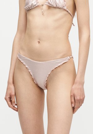 Kobieta w jasnoróżowych, teksturowanych dołach od bikini i dopasowanym topie ze złotym zapięciem, stojąca na jednolitym białym tle.