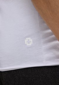 Close-up van de zoom van een wit shirt met een klein wit kruislogo bovenop zwarte denim jeans en een deel van een arm.