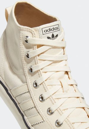 Gros plan d'une basket montante crème Adidas en toile, avec des œillets métalliques, des lacets blancs et le logo Adidas sur la languette.