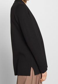 Svart stickad blazer med en avslappnad passform, sjalkrage och sidsprund. Har långa ärmar och en slät yta.