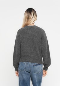 Frau mit langen blonden Haaren, die einen dunkelgrauen Pullover und blaue Jeans trägt, mit dem Rücken zu einer einfarbigen weißen Wand.