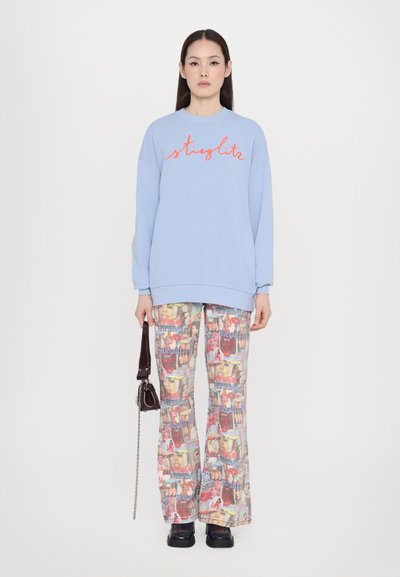 Light blue sweatshirt with red cursive text, paired with patterned wide-leg pants featuring multicolored graphic images. Dark handbag and shoes.