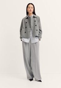 Grauer Wollmischung-Doppelknotenmantel mit schwarzen Knöpfen, getragen über einem hellblau gestreiften Button-Down-Hemd und grauen weiten Hosen.