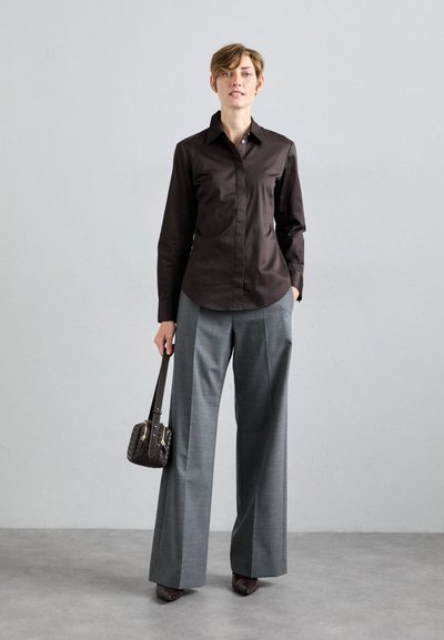 Persona che indossa una camicia a maniche lunghe marrone scuro, pantaloni grigi a gamba larga, mentre tiene una piccola borsa a tracolla intrecciata scura, in piedi su un pavimento grigio.
