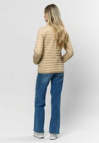 Beige pūkainu jaka ar izšuvumu, garām piedurknēm un augstu apkakli, apvienota ar ziliem džinsiem un baltām sporta apaviem. Skats no aizmugures.