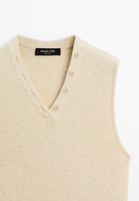 Maglione senza maniche beige a costine con scollatura a V, dotato di sette bottoni decorativi sulla pattina e una texture morbida. Etichetta del marchio visibile.
