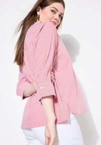 Blusa a righe rosse e bianche con una vestibilità rilassata, maniche arrotolabili e fessure laterali; realizzata in tessuto leggero con una texture liscia.