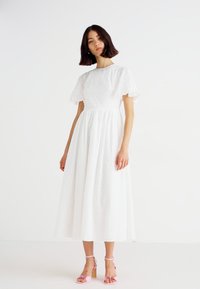 Witte midi-jurk met korte, gerimpelde mouwen en een textuurstof, getailleerde taille, vloeiende rok, gecombineerd met roze strappy hakken.