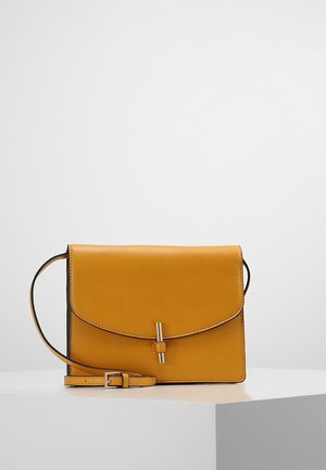 Okergele leren crossbodytas met klepsluiting en goudkleurig verticale stangdetail, geplaatst op een wit geometrisch oppervlak.