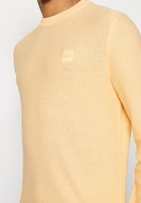 Ljusbeige stickad tröja med rund halsringning och liten fyrkantig "BOSS" logopatch på vänster bröst, bärs av en person.