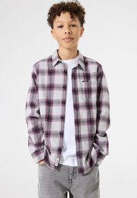 Jongen met krullend haar die een paars en grijs geruit overhemd over een wit T-shirt draagt, met zijn handen in de zakken van grijze broek tegen een effen achtergrond.
