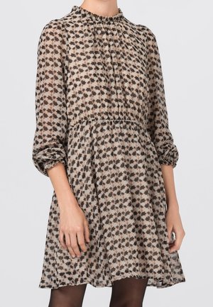 Kobieta ubrana w beżową sukienkę z czarnym kwiatowym wzorem, długimi przezroczystymi bufkami na rękawach, marszczoną talią i falbankowym, wysokim kołnierzem.