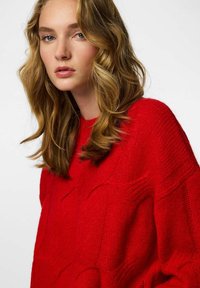 Czerwony sweter dziergany na drutach z teksturowanym, geometrycznym wzorem i luźnym krojem. Długie, lekko opuszczone rękawy oraz okrągły dekolt.