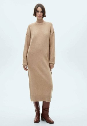 Beige knitted dress with long sleeves, featuring a high neck and ribbed cuffs. Paired with dark brown knee-length boots.