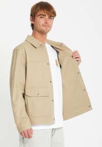 Chaqueta de lona beige con cuello, cierres de botones a presión y dos bolsillos frontales. Llevada abierta sobre una camisa blanca.